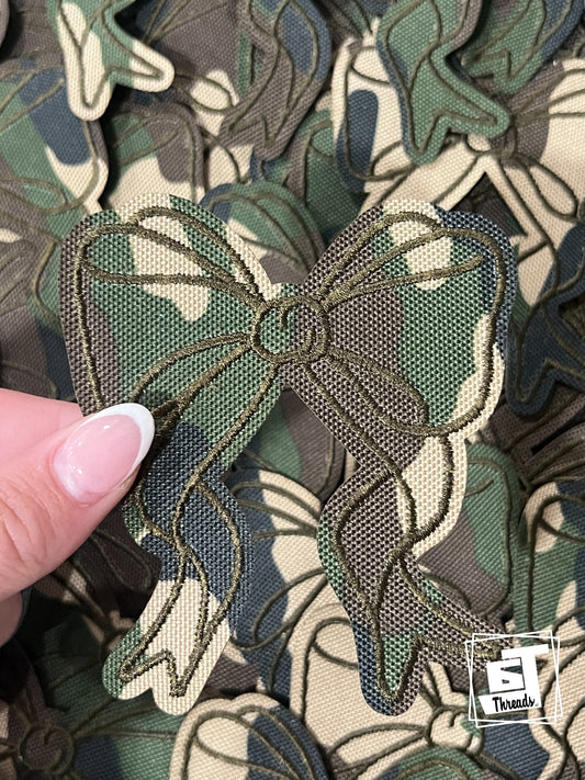 Camouflage-patterned fabric with a bow design held by a hand, branded 'S Threads'.