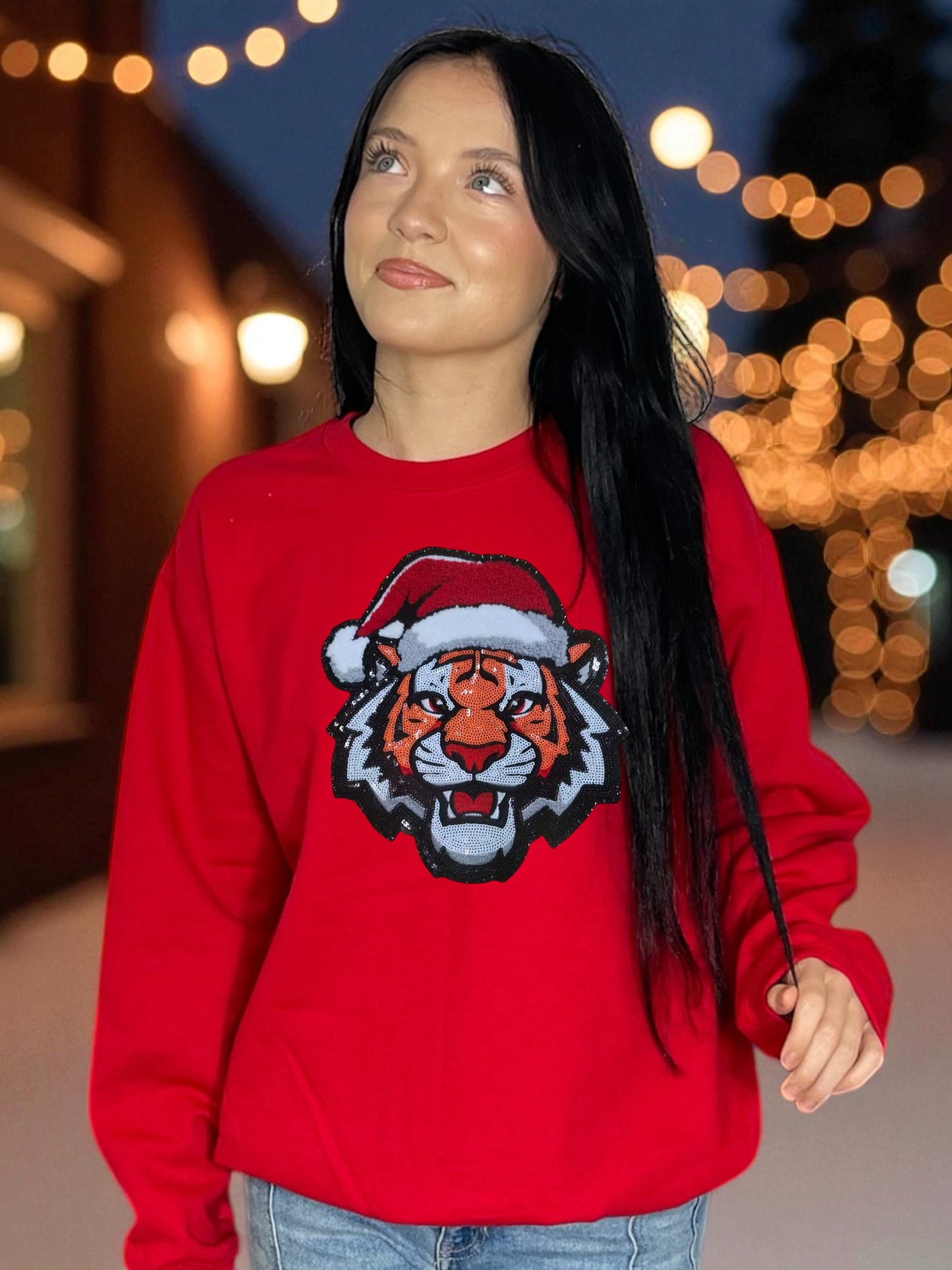 Person wearing a red sweater with a tiger design and Santa hat sequin patch design, standing outdoors with blurred lights in the background.