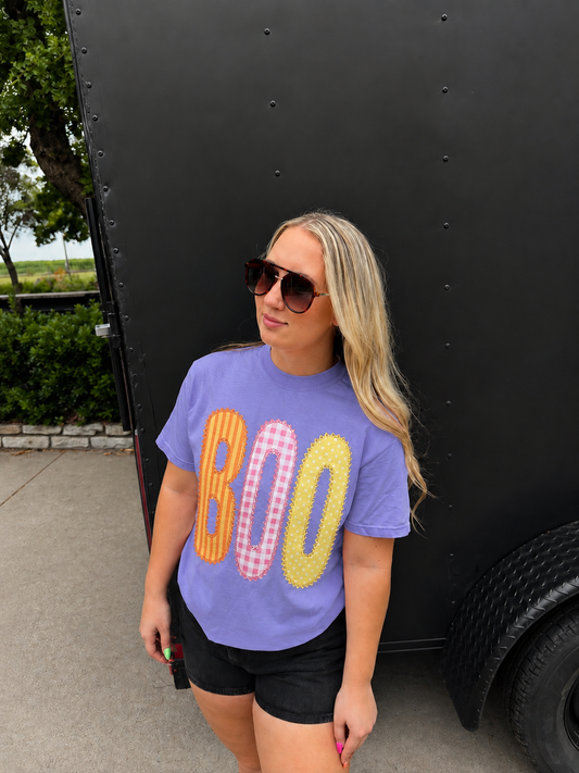 A woman wearing a lavender purple graphic t-shirt with 'BOO' in orange, pink-and-white gingham, and yellow gingham lettering, standing outdoors next to a black trailer.