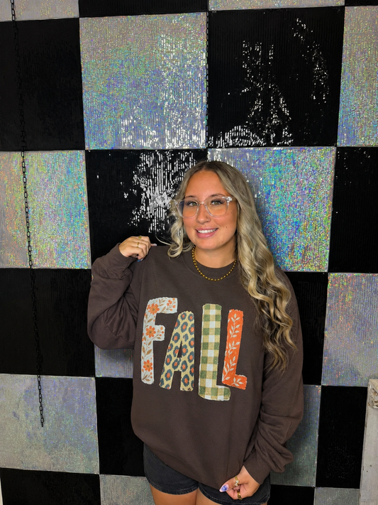 A woman with glasses and long blonde hair stands in front of a black and iridescent checkered wall, wearing a brown sweatshirt with the word 'FALL' spelled out in decorative patterned letters.