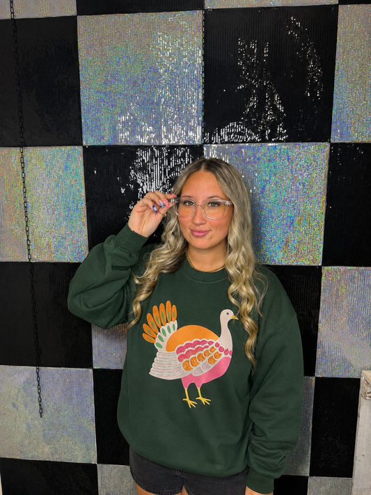 A woman with blonde wavy hair and glasses wearing a dark green crewneck sweatshirt with a colorful turkey graphic, standing in front of a black and holographic checkered wall.