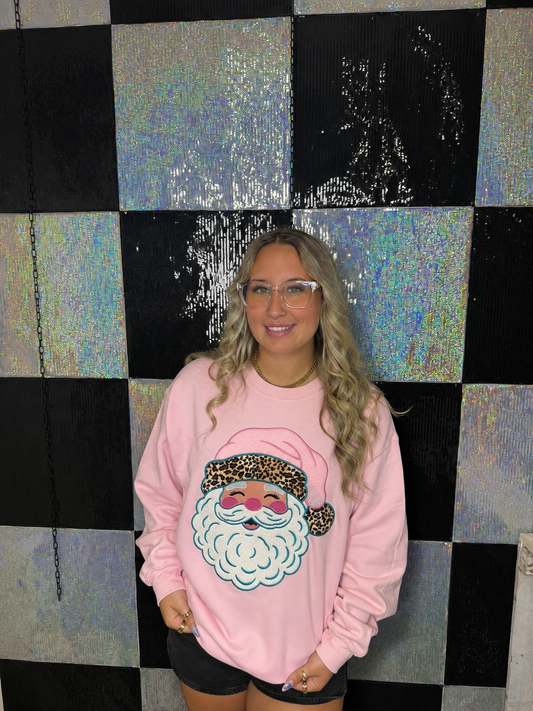 A woman with blonde hair and glasses wearing a pink crewneck sweatshirt with a sequined Santa face graphic, standing in front of a black and iridescent checkered wall.