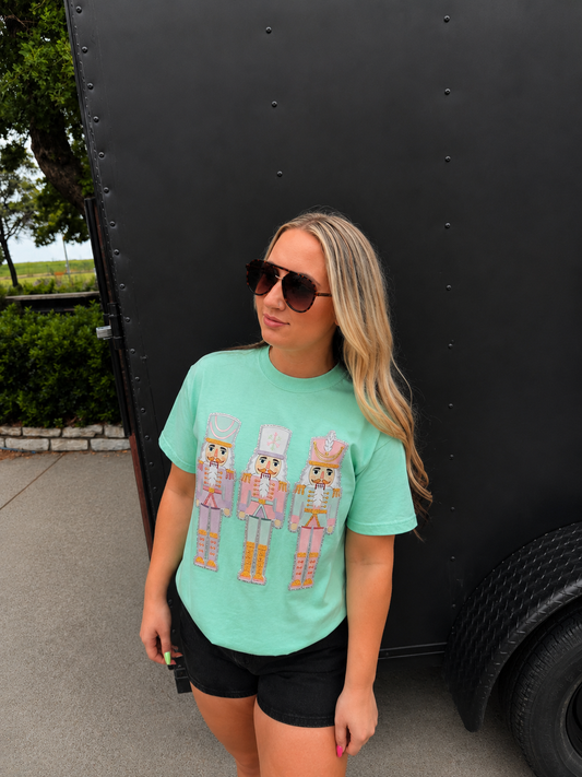 A woman wearing a mint green graphic t-shirt with three nutcracker illustrations, black shorts, and sunglasses, standing outdoors next to a large black trailer.