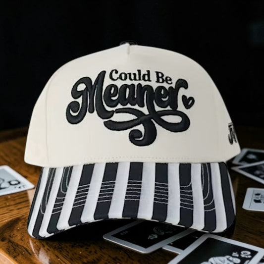 White cap with black text 'Could Be Meaner' on a wooden surface with playing cards.