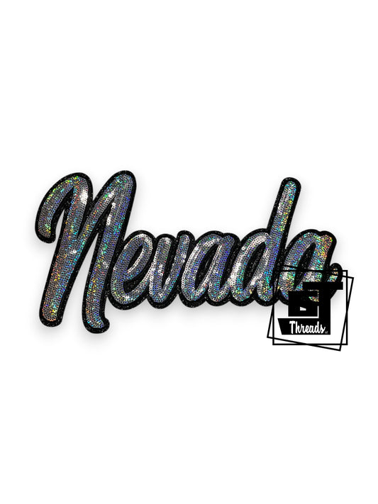 This iron-on patch features a striking silver cursive "Nevada" design adorned with shimmering sequins. Black embroidery details and a glitter-infused background create depth and visual interest. Measuring 12 inches wide by 7 inches tall, this decorative patch is sized for maximum impact on jackets, bags, or apparel projects.