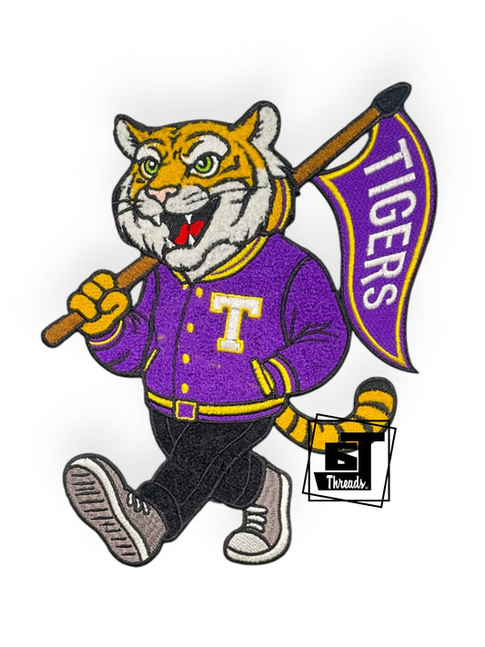 Cartoon tiger mascot holding a flag with 'Tigers' text, wearing a purple jacket with a 'T' logo.