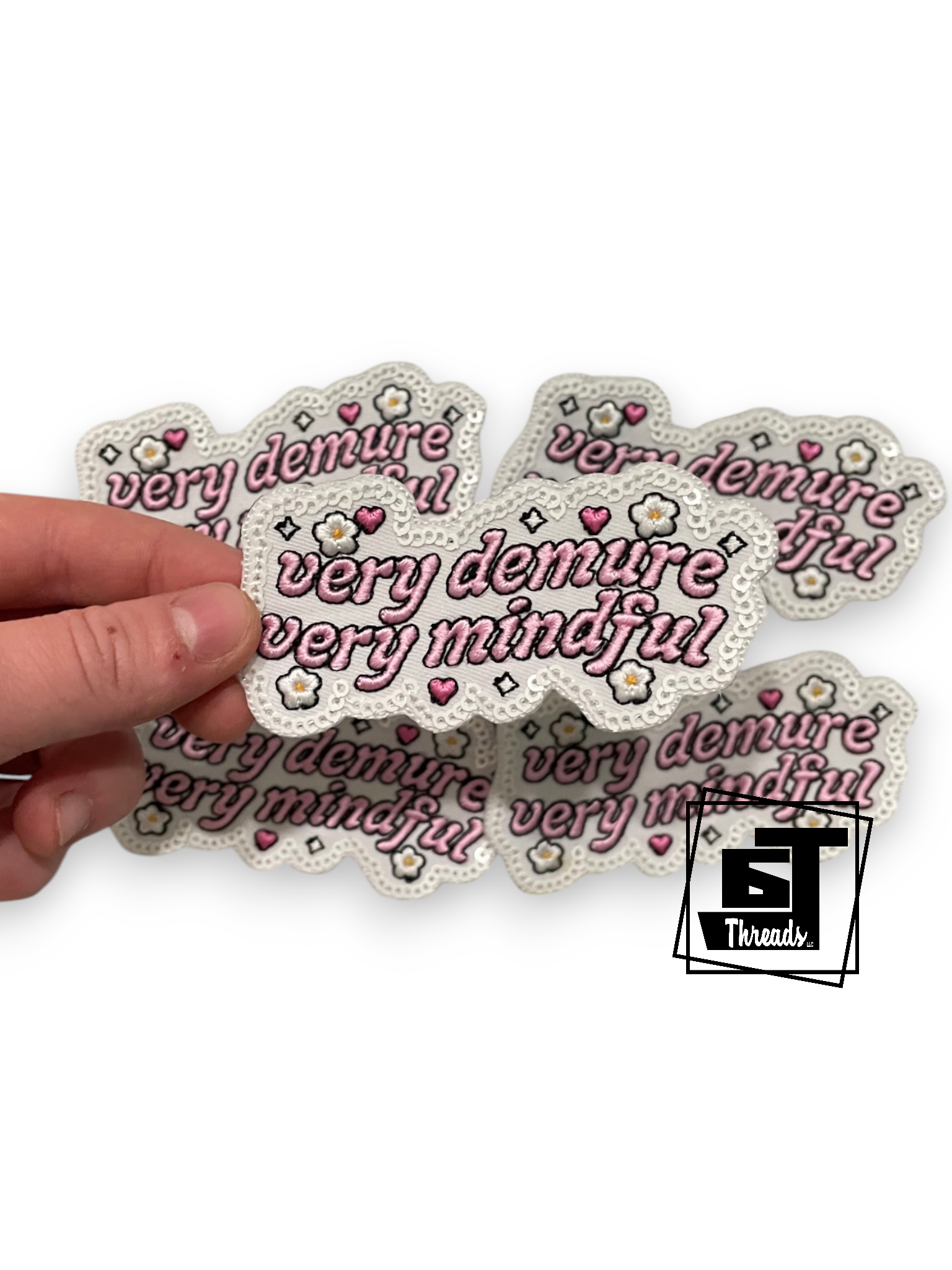 A hand holding a stack of white scalloped patches with the text 'very demure and very mindful' in pink script, decorated with small pink hearts, white flowers, and pink bows. A '5 Threads' logo is visible in the bottom right corner.