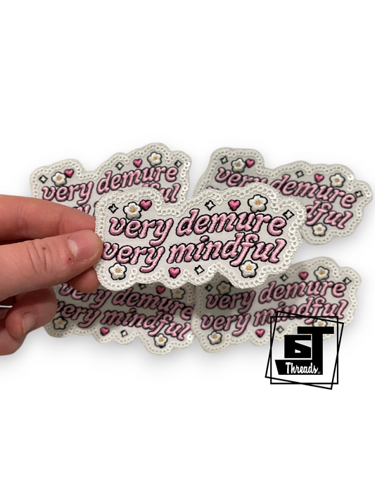 A hand holding a stack of white scalloped patches with the text 'very demure and very mindful' in pink script, decorated with small pink hearts, white flowers, and pink bows. A '5 Threads' logo is visible in the bottom right corner.