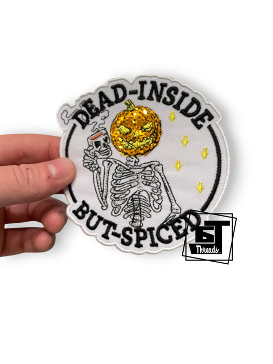 A hand holds a circular embroidered patch featuring a skeleton and a glittery gold jack-o'-lantern head, with the text 'DEAD-INSIDE BUT-SPICED' on a white background. A 'BT Threads' logo watermark is visible in the lower right corner.