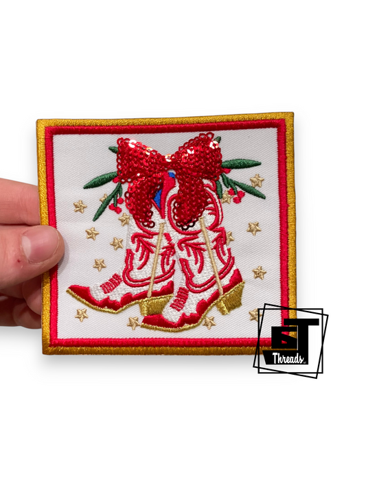 An embroidered patch featuring a pair of red cowboy boots with a large red bow, green leaves, and gold stars on a white background, held by a hand on the left. A 'ST Threads' logo is visible in the bottom right corner.