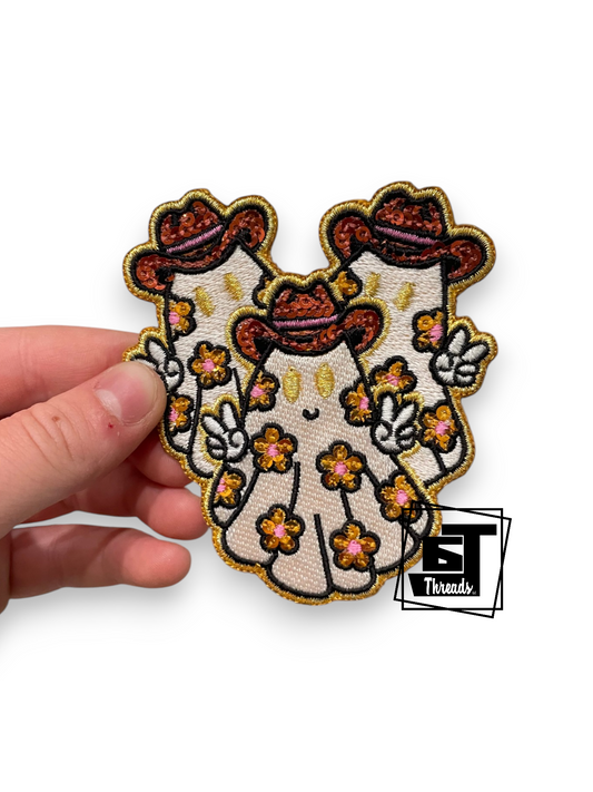 An embroidered patch shaped like a stylized Santa Claus figure in a white suit with orange and yellow floral details, held by a hand against a white background. A 'BT Threads' logo is visible in the bottom right corner.