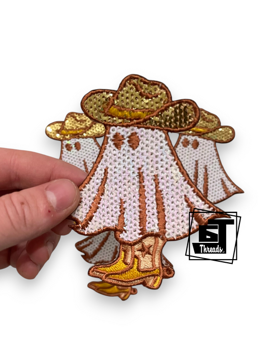 A hand holding a small embroidered patch featuring a ghostly figure in a white dress, gold cowboy hats, and yellow boots, with a black and white 'ST Threads' logo watermark in the lower right corner.