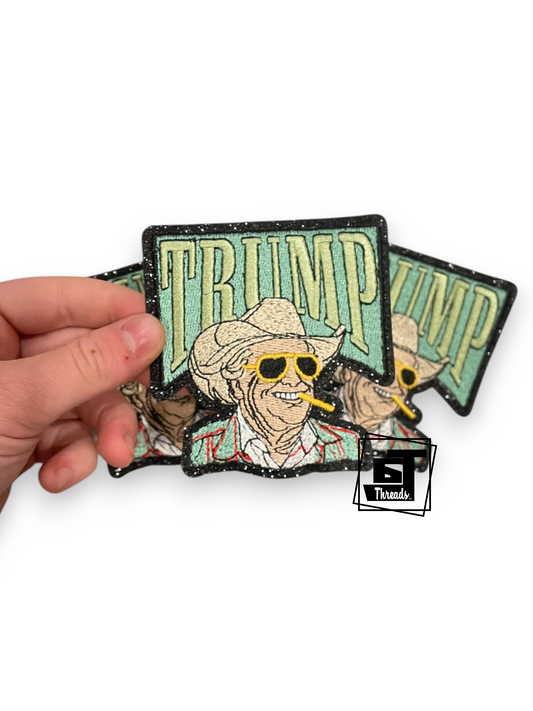 A hand holds three embroidered patches featuring a stylized cowboy figure with a hat, sunglasses, and a cigar, with the word 'TRUMP' in large letters above. The patches have a distressed, grunge aesthetic with black outlines and speckled details, set against a plain white background.