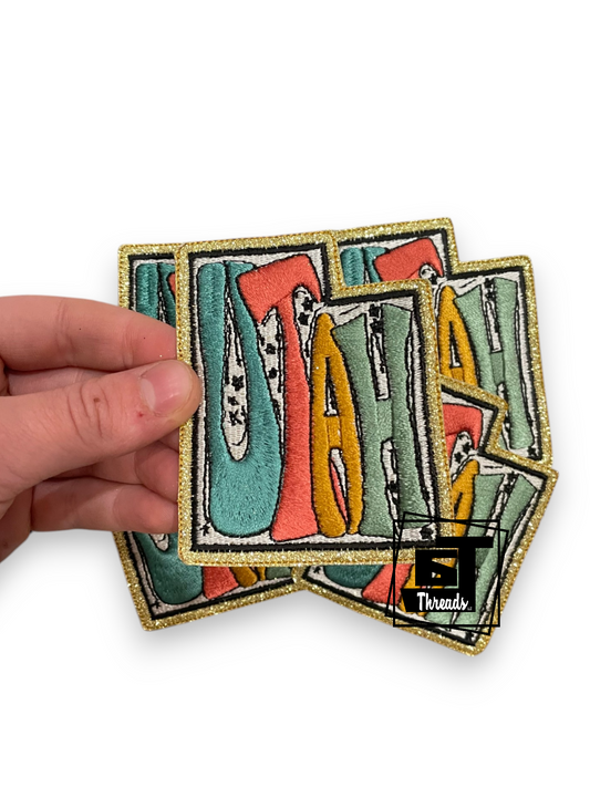 A hand holds a stack of embroidered patches featuring stylized letters spelling 'JAZZ' in teal, coral, mustard yellow, and sage green with a glittery gold border, on a white background.