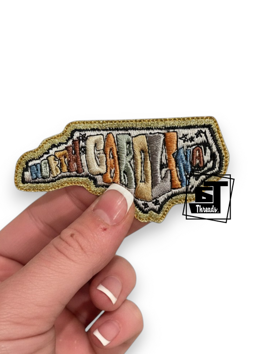 A hand holding a small embroidered patch shaped like the state of New Hampshire, with the word "NORTH HANPEMA" stitched across it in multicolored letters. A black and white "ET Threads" logo watermark is visible on the right side of the patch.
