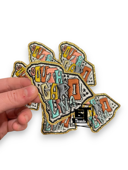 A hand holding three embroidered patches shaped like a fish, featuring colorful graffiti-style lettering that reads 'South Carolina' with gold glitter borders on a white background.
