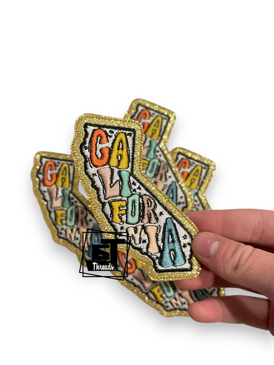A hand holding a fanned-out set of embroidered California state patches with a glittery gold border and multicolored lettering, on a white background.