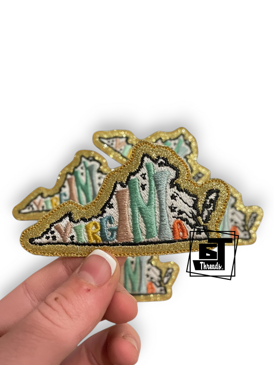 A hand holding a glittery embroidered patch shaped like the state of Nevada, featuring a mountain design with pastel green, blue, and beige details and a gold glitter border, on a white background.