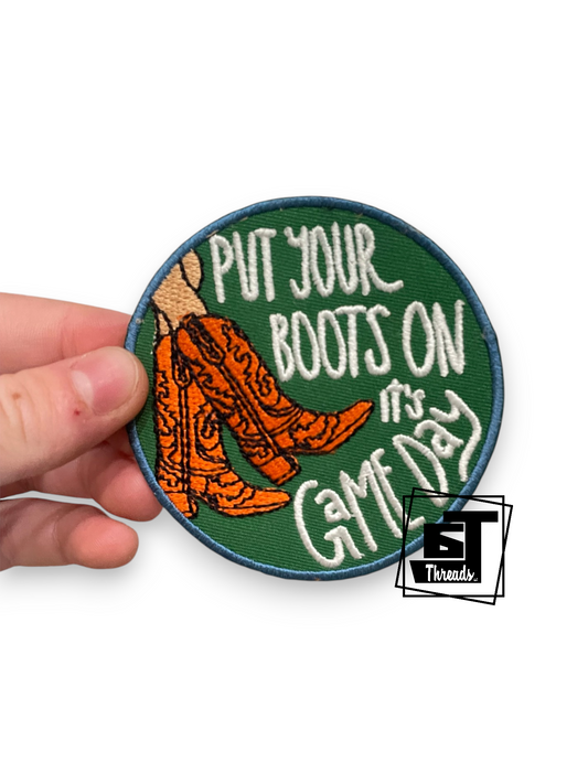 A hand holds a round embroidered patch featuring the phrase 'PUT YOUR BOOTS ON it's GATE DAY' in white lettering over a green background, with orange cowboy boots and a tan boot on the left. A small 'BT Brands' logo appears in the lower right corner.