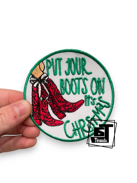 A circular embroidered patch held by a hand against a white background. The patch features green text reading 'PUT JOUR BOOTS ON it's CHRISTMAS' with an illustration of red boots and a beige hat.
