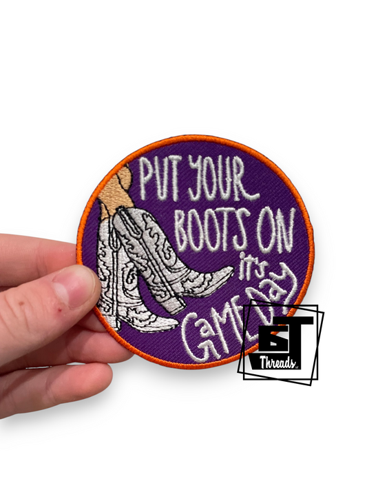 A hand holding a round embroidered patch with a purple background, orange border, white text reading 'PUT YOUR BOOTS ON in Game Day,' and a white boot illustration. A small 'BT Threads' logo watermark appears near the lower right.