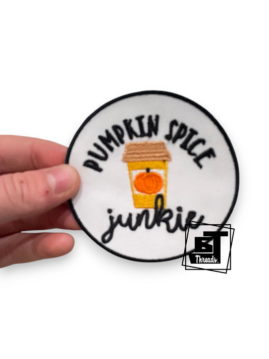 A hand holding a round embroidered patch that reads 'PUMPKIN SPICE junkie' with a small pumpkin coffee cup illustration in the center, on a white background.