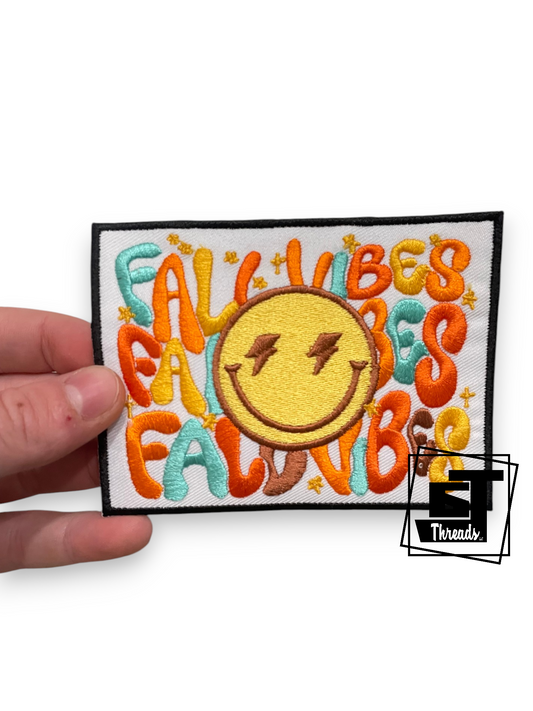 An embroidered patch held by a hand, featuring a yellow smiley face with lightning eyes in the center, surrounded by colorful bubble letters spelling 'FALLO VIBES FALLO NIB' on a white background with a black stitched border.