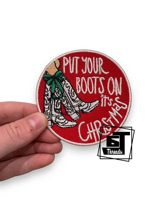A hand holding a round embroidered patch with a red background, white text reading 'PUT YOUR BOOTS ON IT'S CHRISTMAS,' and an illustration of white boots with a green bow. A 'BT Threads' logo watermark is visible in the lower right.