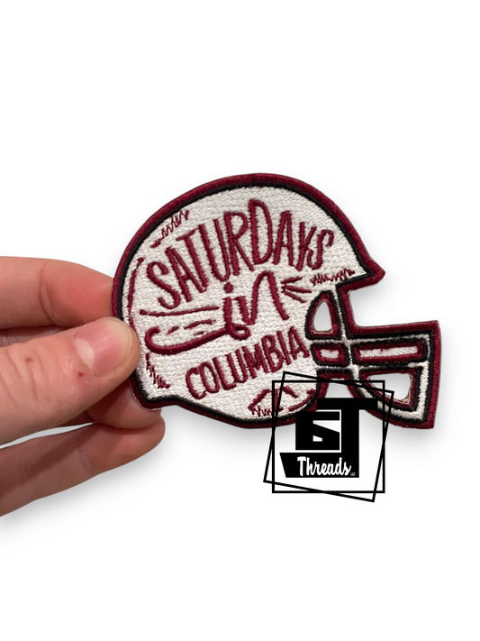 A hand holding an embroidered football helmet patch with the text 'SATURDAY IN COLUMBIA' in burgundy on a white background. A '5 Threads' logo watermark is visible in the lower right.