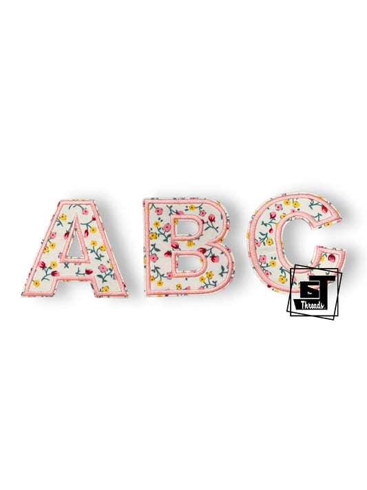 Three decorative capital letters A, B, and C with a pink floral pattern, displayed on a clean white background. A small black logo for 'St.