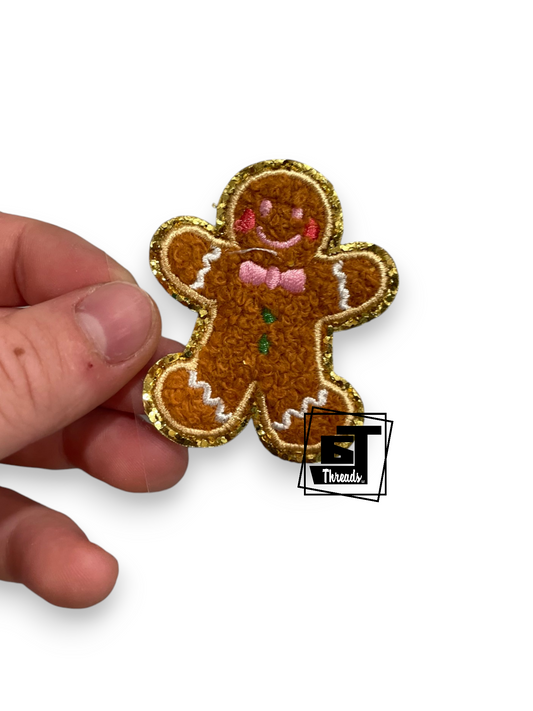 A hand holding a small gingerbread man patch against a plain white background. The patch is brown with a glittery gold border, pink embroidered cheeks and bow tie, and white piping details.
