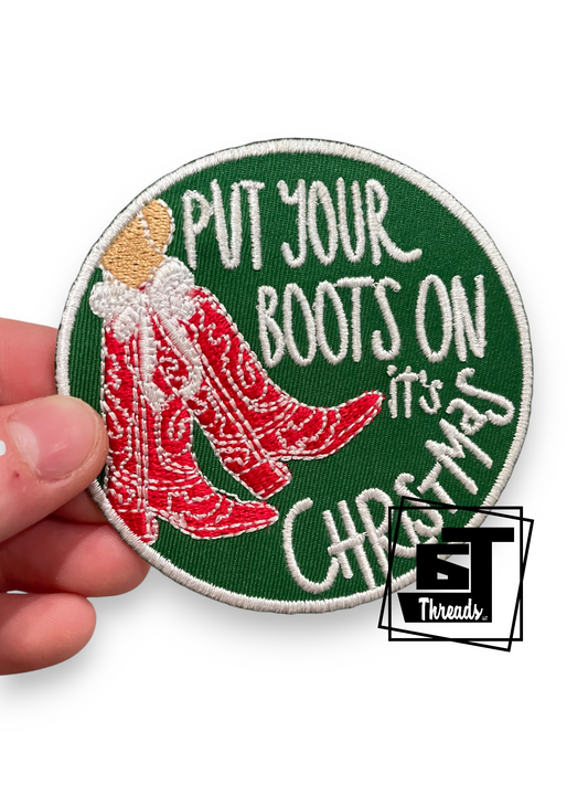 A hand holding a round embroidered patch with a green background, white border, red cowboy boots, and white text reading 'PUT YOUR BOOTS ON it's CHRISTMAS'. A small 'BT Threads' logo watermark is visible in the lower right corner.