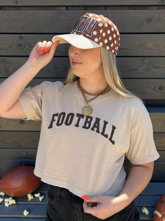 Person wearing a 'FOOTBALL' t-shirt and polka dot cap, standing against a wooden background with sports balls and popcorn.