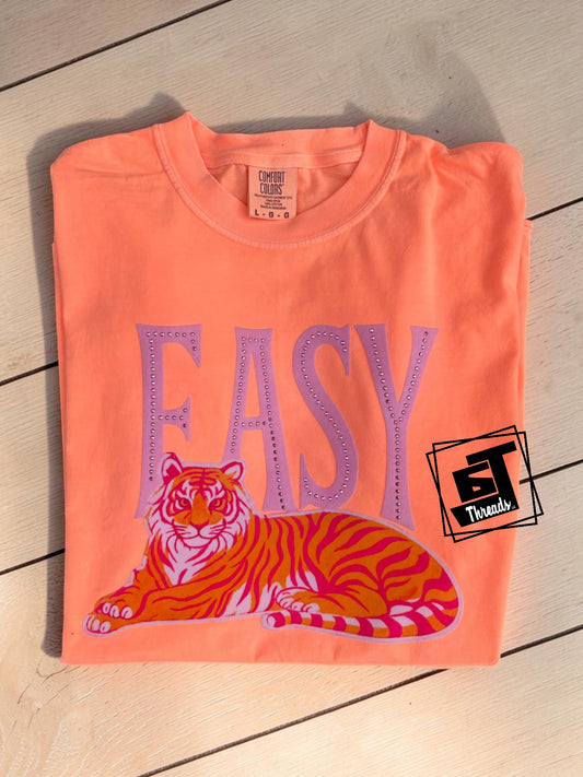 Folded orange t-shirt with a tiger graphic and 'EASY' text on a wooden surface