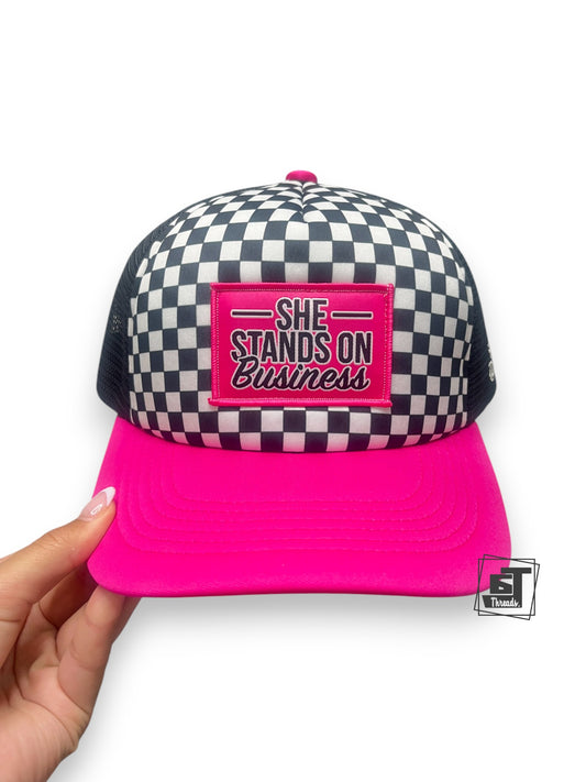 Checkered cap with pink brim and 'She Stands on Business' patch held by a hand on a white background