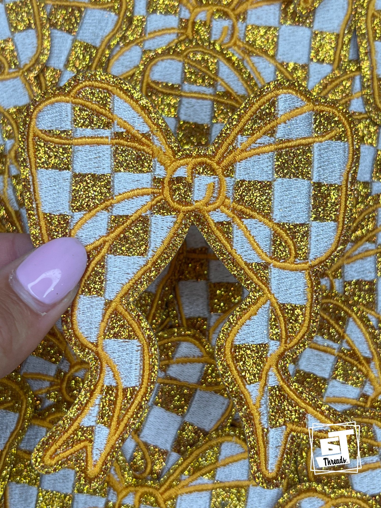 Gold embroidered fabric with a checkered pattern on a light blue background, held by a hand.