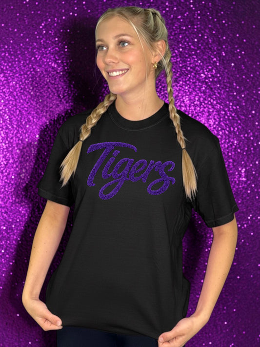 Person wearing a black t-shirt with 'Tigers' in purple text against a purple glittery background