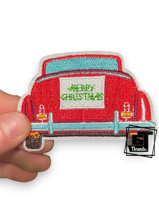 Embroidered patch of a red truck with 'Merry Christmas' text held by a hand on a white background.