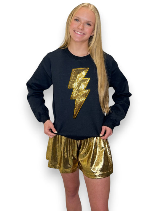 Young girl wearing a black sweatshirt with a gold lightning bolt and gold sequin shorts on a white background