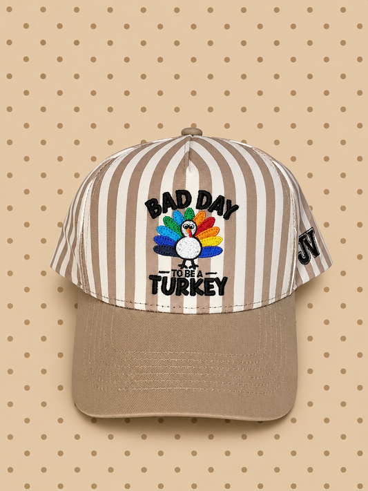 A baseball cap with beige and white vertical stripes, featuring a brown visor. The front has an embroidered design of a turkey with a rainbow tail feathers and the text 'BAD DAY TO BE A TURKEY' in black.