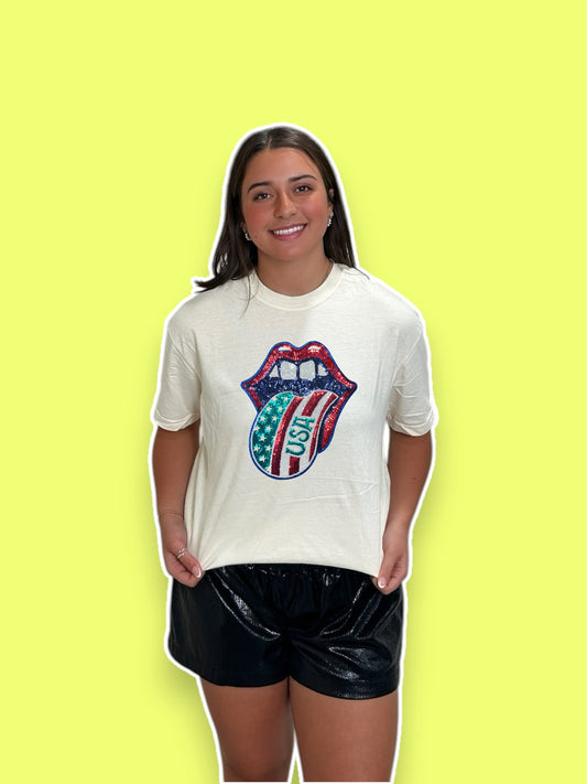A person wearing a white t-shirt with a sequin design in the shape of a tongue sticking out, with red, blue, and white colors, resembling the American flag.