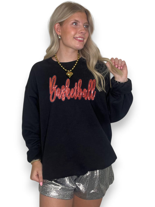 Women with blonde hair wearing a black crewneck with an orange sequin cursive basketball and silver shorts with white background.
