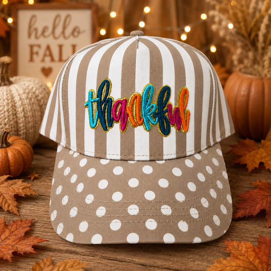 A baseball cap styled for autumn, featuring vertical brown and white stripes on the crown with colorful embroidered 'thankful' text, and a brown brim with white polka dots. The cap is set on a rustic wooden surface surrounded by fall leaves, pumpkins, and a warm, cozy background.